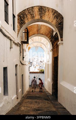 Deux touristes marchant sous des arches historiques en briques le long d'une étroite rue pavée bordée de bâtiments blanchis à la chaux à Vejer de la Frontera, Cádiz, Anda Banque D'Images