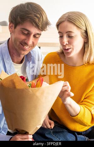 Jeune couple souriant en partageant un bouquet surprise de tulipes et de roses à la maison, profitant d'un tendre moment de romance, de gratitude et de célébration Banque D'Images