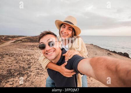 Jeune homme donnant un tour sur ses épaules à sa petite amie tout en souriant à l'appareil, capturant un selfie joyeux ensemble pendant leurs vacances au bord de l'océan au coucher du soleil Banque D'Images