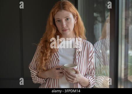 Jeune femme rousse tenant une tasse et regardant pensivement par la fenêtre, portant une chemise rayée dans un portrait intérieur calme du matin Banque D'Images
