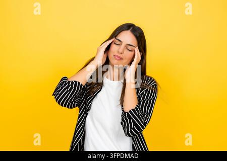 Jeune femme souffrant d'un mal de tête sévère, tenant ses deux mains contre ses tempes et fermant les yeux avec une expression de douleur, debout devant un fond jaune vif Banque D'Images