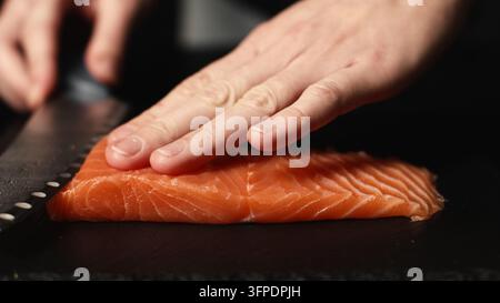 Mains d'un chef professionnel coupant un filet de saumon cru frais pour la préparation de sushi sur une planche à découper sombre Banque D'Images