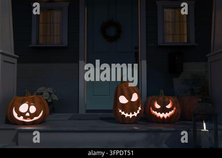 Des jack-o'-lanterns lumineux et effrayants accueillent les invités. Trois citrouilles sculptées illuminées arborant des visages distincts et variés ornent un porche sombre en banlieue près d'une porte bleue Banque D'Images