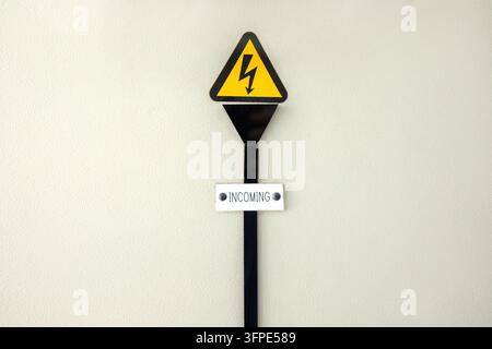 Panneau d'avertissement de haute tension avec un éclair sur un triangle jaune au-dessus de l'étiquette "INCOMING" sur un poteau noir, monté sur un mur beige, symbole de danger électrique Banque D'Images