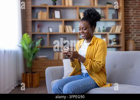 Femme afro-américaine souriante assise confortablement sur un canapé, parcourant du contenu et communiquant sur son téléphone portable, profitant de la technologie sans fil et de l'accès à Internet Banque D'Images