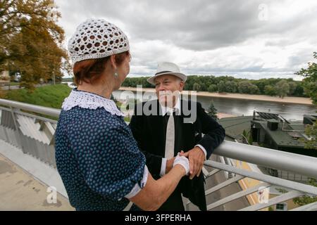 Un couple vêtu d'une tenue vintage profite d'un moment tendre ensemble sur une promenade pittoresque surplombant une rivière, entouré d'une végétation luxuriante et d'un ski nuageux Banque D'Images