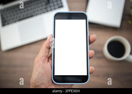 Main tenant un smartphone avec écran blanc vierge au-dessus d'un espace de travail extérieur avec un ordinateur portable, du café et un cahier en arrière-plan flou. Application mobile et site web Banque D'Images