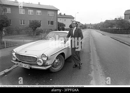 Un moment figé dans le temps : une Volvo P1800 des années 1960 et son propriétaire mystérieux Cette photographie nostalgique capture une Volvo P1800 classique des années 1970, avec son fier propriétaire posant à ses côtés. Équipée d’un moteur à essence de 76 kW (103 ch) et d’une boîte manuelle, cette voiture de sport suédoise emblématique représente le sommet du design vintage de Volvo.  Une plongée approfondie dans le registre des véhicules révèle une chronologie fascinante :    Le lancement : La voiture a été enregistrée pour la première fois le 11 octobre 1967 en tant que modèle de 1968.    Hors des radars : Étonnamment, la voiture est officiellement hors d’usage (SORN) depuis 1981, ce qui signifie qu’elle a passé plus de quatre Banque D'Images