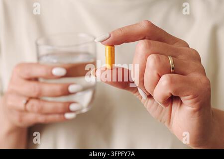 Femme tenant une capsule de supplément jaune près d'un verre d'eau. Contexte de soins de santé pour les vitamines orales, les médicaments quotidiens, la nutrition et la routine de bien-être Banque D'Images