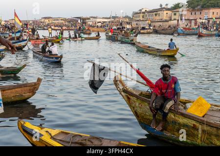 Un jeune homme est assis à la proue d'un bateau de pêche en bois dans le port, région centrale, Elmina, Ghana Banque D'Images