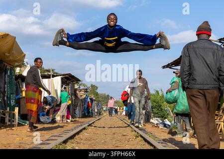 Jeune danseur masculin vêtu d'une veste en velours bleu exécutant un saut fendu en plein air au-dessus des voies ferrées au ballet de Kibera, comté de Nairobi, Nairobi, Kenya Banque D'Images