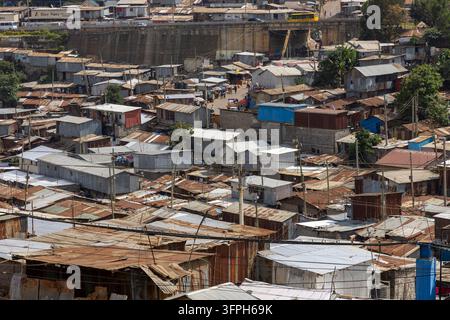 Vue sur les toits densément recouverts de tôle ondulée du quartier de Kibera avec une rue animée et un bus jaune traversant un pont en arrière-plan, Comté de Nairobi Banque D'Images