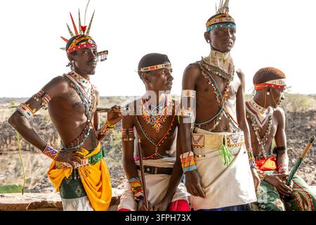 Quatre hommes de la tribu Samburu avec des colliers de perles et des coiffes à plumes posant ensemble sur un mur de pierre au-dessus d'une gorge rocheuse dans le paysage sec sous un ciel lumineux Banque D'Images
