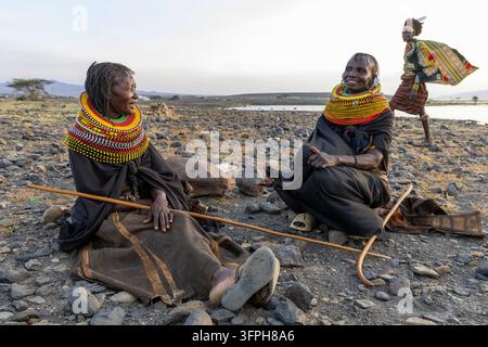 Deux femmes de la tribu Turkana assises et riant sur un sol pierreux tandis qu'un homme vêtu d'une jupe imprimée et de plumes marche derrière elles près du lac Turkana, Marsabit Banque D'Images