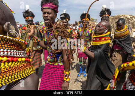Homme de la tribu Turkana portant une chemise colorée imprimée dansant avec un fouet tressé à côté d'une femme âgée en cape noire au festival culturel du lac Turkana Banque D'Images