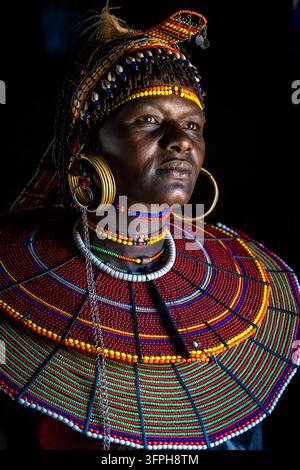 Portrait dramatique d'une femme de la tribu Pokot sur fond noir avec un coiffelet à plumes, des colliers de coquillages, des boucles d'oreilles en laiton et un large collier rouge et vert Banque D'Images