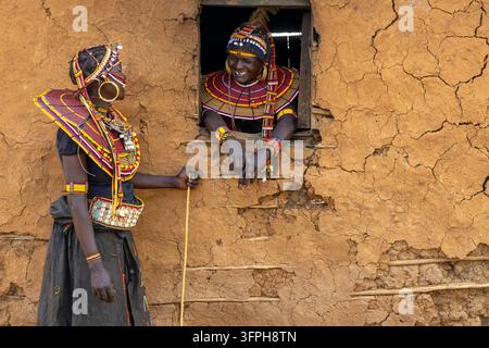 Deux femmes de la tribu Pokot riant ensemble, l'une tenant un bâton à côté d'un mur de boue et l'autre se penchant par une petite fenêtre avec des colliers de perles, B Banque D'Images