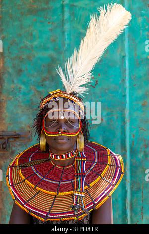 Portrait d'une femme du tribe Pokot avec une grande plume d'autruche blanche dans son bandeau, debout devant une porte en métal turquoise rouillée, portant un grand collier de perles Banque D'Images