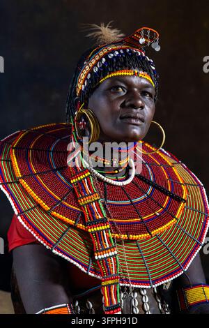 Portrait d'une femme de la tribu Pokot dans une hutte sombre portant un coiffeur traditionnel en plumes, des colliers de coquilles de cauri, des boucles d'oreilles en laiton et un énorme collier de perles, regardant vers le haut Banque D'Images