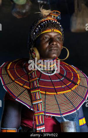 Femme de la tribu Pokot dans une hutte en boue sombre avec un couvre-chef à plumes, un bandeau en coquillage, des boucles d'oreilles en laiton et un large collier de perles rouges et vertes, regardant sur le côté, B Banque D'Images