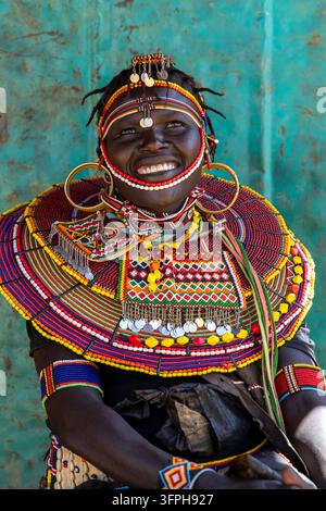 Femme souriante de la tribu Pokot regardant vers le haut devant une porte métallique turquoise rouillée, portant un collier de perles, des boucles d'oreilles en laiton et des disques en argent sur la tête Banque D'Images