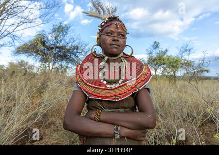 Femme du tribe Pokot avec un bandeau à plumes et coquilles de cowrie et un large collier de perles rouges, debout dans la brousse sèche les bras croisés, Narok, Chepilat, Kenya Banque D'Images