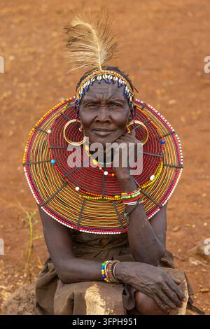 Femme du tribe Pokot avec un bandeau à plumes et coquilles de cauri et un large collier de perles, assise sur un rocher dans le sol rouge, comté de Narok, Chepilat, Kenya Banque D'Images