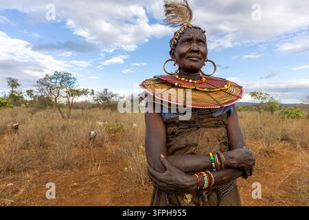 Femme de la tribu Pokot avec un bandeau à plumes et à coquilles et un large collier de perles, debout dans la brousse sèche les bras croisés, Narok, Chepilat, Kenya Banque D'Images