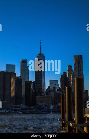 One World Trade Center et les gratte-ciel de Lower Manhattan vus à travers l'eau à New York City, avec les pilotis sous un ciel bleu clair. Banque D'Images