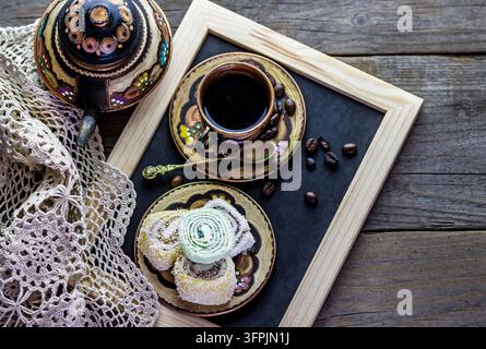 Vue aérienne d'un café noir dans une tasse en céramique décorative avec des grains de café, une cuillère et des sucreries en poudre à côté d'un pot peint sur une table en bois. Banque D'Images
