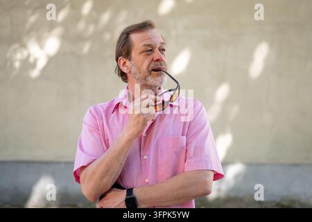 Un homme d'âge moyen en chemise rose tient des lunettes de soleil tout en regardant sur le côté à l'extérieur. Banque D'Images