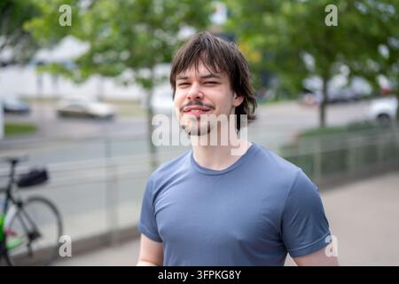 Un jeune homme souriant se tient en plein air avec une rue et des arbres légèrement flous en arrière-plan. Banque D'Images