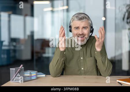 Représentant du service client masculin portant un casque, assis à un bureau, se sentant submergé et frustré pendant qu'il communique avec un client difficile ou gère un problème technique Banque D'Images