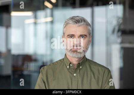 Homme d'affaires mûr aux cheveux gris et à la barbe portant une chemise décontractée, debout avec assurance dans un environnement de bureau moderne, incarnant le professionnalisme et l'expérience Banque D'Images