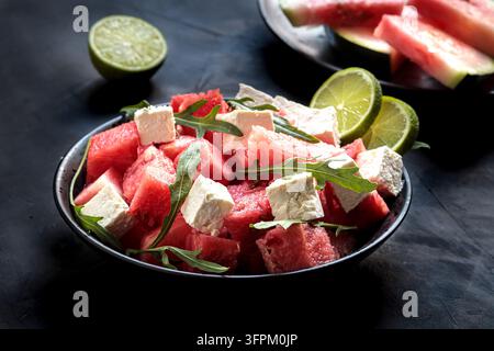 Salade d'été fraîche aux cubes d'eau-de-melon juteux, au fromage feta crémeux et aux feuilles d'arugula vertes dans un bol noir, servie avec des moitiés de citron vert. Plat léger et sain f Banque D'Images