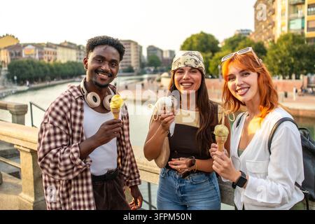 Trois amis souriants et profitant d'un gelato ensemble par une journée d'été ensoleillée le long des canaux Navigli de Milan, explorant le front de mer, le paysage urbain et l'insouciance Banque D'Images