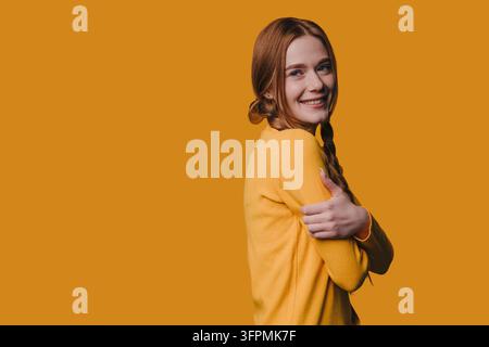 Jeune femme rousse souriant par-dessus son épaule tout en s'embrassant contre un fond orange, portant un pull jaune, portrait de studio montrant des tresses Banque D'Images