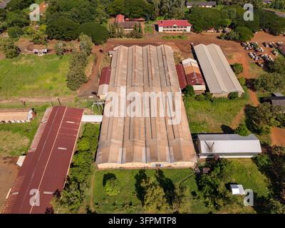 Vue aérienne de la plantation de caoutchouc Chup présentant une grande installation industrielle avec un toit en tôle ondulée entourée de végétation luxuriante et de routes en terre dans Banque D'Images