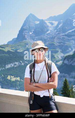 Randonneuse avec sac à dos, chapeau de soleil et lunettes de soleil miroir posant les bras croisés devant le paysage montagneux de l'Eiger lors d'un voyage estival ensoleillé à Murren, Alpes suisses Banque D'Images
