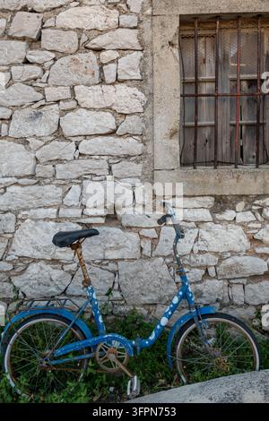 Vieux vélo pliant bleu appuyé contre un mur de pierre usé sous une fenêtre grillagée dans un cadre rustique calme d'un village. Banque D'Images