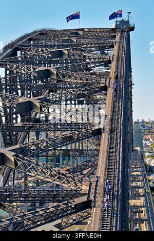 Groupe de touristes en combinaisons d'escalade bleues marchant sur l'arche en acier au-dessus de la circulation routière et de la passerelle piétonne à Sydney, Nouvelle-Galles du Sud, par un après-midi clair. Banque D'Images