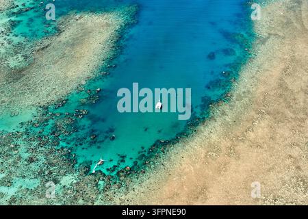 Vue aérienne d'un voilier à l'ancre dans un canal bleu profond bordé de platesses de récif corallien peu profondes et d'eau turquoise sous un soleil éclatant Banque D'Images