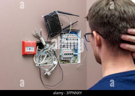 Homme à lunettes face à une boîte de circuit ouverte avec des fils emmêlés, un routeur et une alarme incendie sur un mur rose, symbolisant les complexités technologiques modernes. Banque D'Images