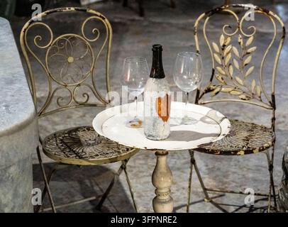 Table romantique en terrasse avec une bouteille de vin fraîche, deux verres vides et des chaises en métal vintage ornées sur un patio en pierre. Banque D'Images