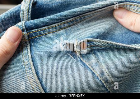 Les mains affichent un trou effiloché à côté du rivet de la poche avant sur un jean bleu et montrent des dommages de denim usés qui nécessitent une réparation minutieuse des vêtements. Banque D'Images