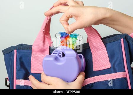 Une main d'enfant place un appareil orthodontique arc-en-ciel et un étui de voyage violet dans un sac pour enfants bleu et rose pour garder l'appareil dentaire propre à l'école. Banque D'Images