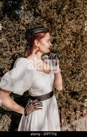 Femme à la mode vintage, dame élégante en robe blanche et gants en dentelle posant devant un fond de verdure sous un soleil d'été éclatant Banque D'Images