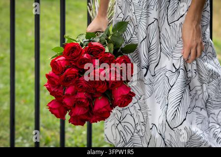 Femme en robe blanche fleurie élégante tenant un bouquet luxuriant de roses rouges éclatantes en plein air, gros plan de fleurs romantiques symbolisant l'amour et la passion sur Banque D'Images