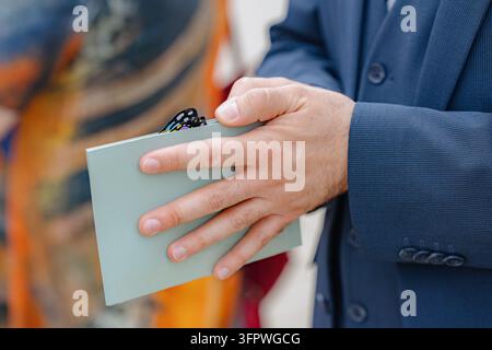 Un homme en costume formel tient un portefeuille à l'intérieur, peut-être lors d'un événement ou d'une réunion. L'image est un plan rapproché se concentrant sur les mains de l'homme et Banque D'Images