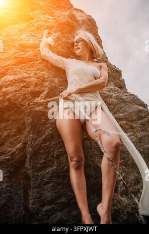 Portrait en bordure de falaise ensoleillé, femme blonde en tenue de resort blanche posant devant un fond rocheux sous la lumière chaude du coucher de soleil Banque D'Images